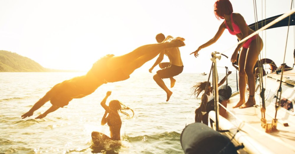 A group of adults jumping off of a boat into the water. One of the people is standing on the boat.