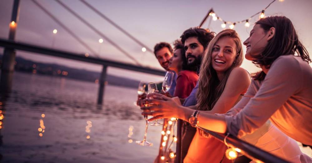 Some friends stand on the side of a boat on the water, smiling and laughing with a lighted bridge in the distance.